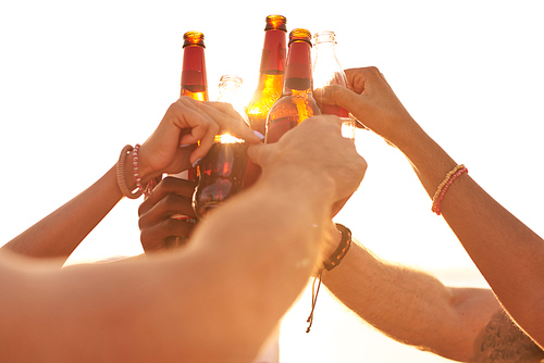 Close-up of unrecognizable friends toasting with beer bottles in summer sunlight while cheering for great open-air party