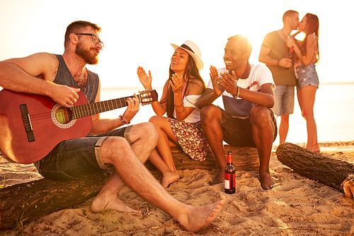 Cheerful talented bearded guy in glasses playing guitar for friends and sitting on log, positive enjoyable young couple supporting him with applause on beach