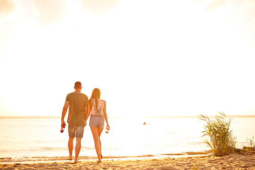 Rear view of affectionate young couple carrying bottles and holding hands while walking on sand and coming to sea water