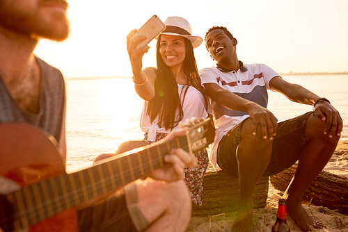 Cheerful beautiful Hispanic girl taking selfie with African boyfriend while they sitting on log and listening to street musician on beach