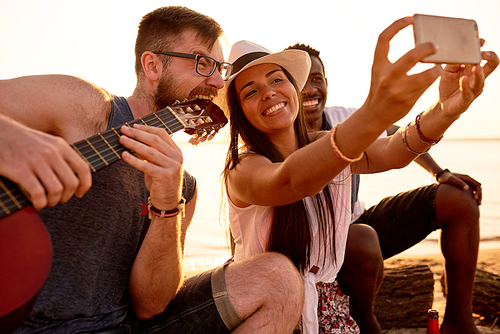Cheerful happy pretty Latin woman in stylish hat and photographing together with African friend and crazy musician in eyeglasses biting guitar for great picture on beach.