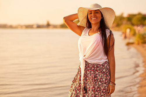 Smiling cheerful beautiful Hispanic woman in summer outfit adjusting sun hat and  while standing alone on beach