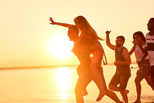 Cheerful excited young man giving piggyback to ecstatic girlfriend waving hands, he running in front of friends group on beach at sunset