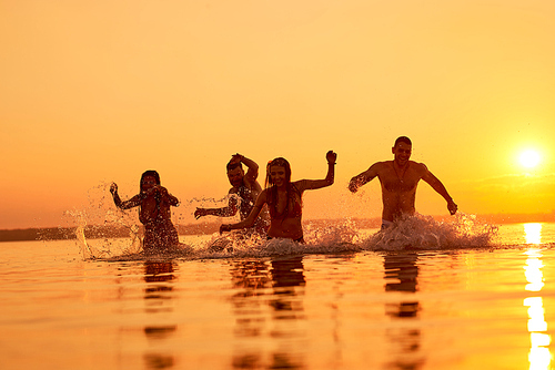 Cheerful emotional young friends running in water while having fun at sunset beach, splashing water around them