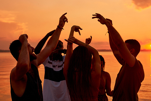 Ecstatic carefree young people dancing with raised hands while having fun at sunset beach