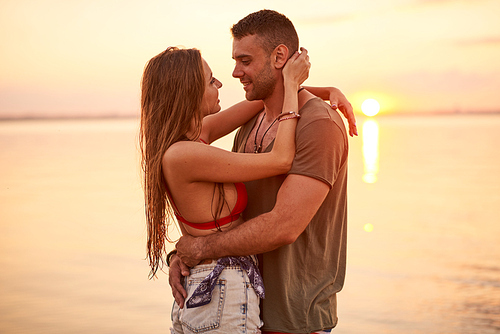 Happy affectionate beautiful young couple in casual clothing hugging each other and looking into eyes during date at sea sunset