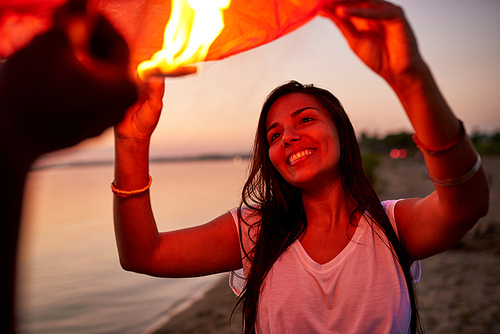 Cheerful attractive young woman with long hair wearing bracelets looking at burning fire inside of Chinese lantern while holding it with boyfriend on beach
