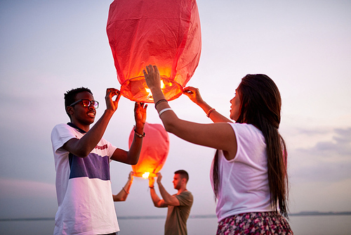 Happy positive young multiethnic couple preparing to launch sky paper lantern while standing on beach, people in background making the same