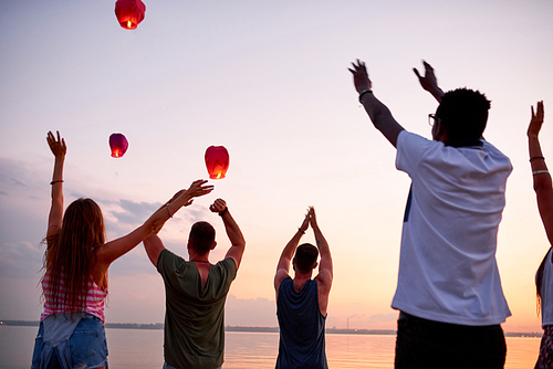 Rear view of excited young people waving hands to flying sky lanterns while standing at sea outdoors