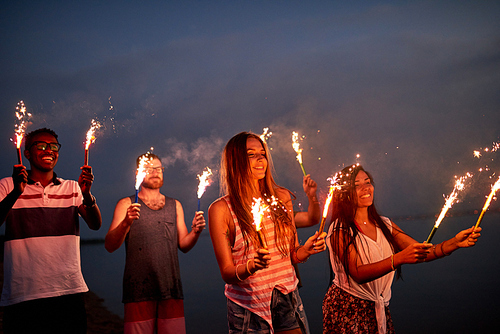 Positive excited young multiethnic friend holding sparklers while having fun at outdoor party and celebrating birthday person
