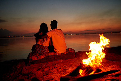 Rear view of romantic couple sitting on log covered with plaid and embracing while contemplating tranquil sea on beach, burning campfire behind them at night