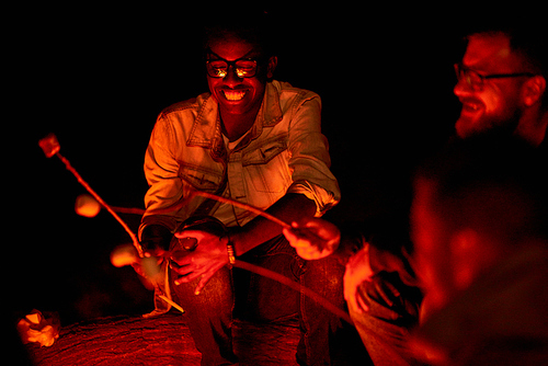Cheerful happy young African man in eyeglasses roasting marshmallow while talking to close friends during night camping