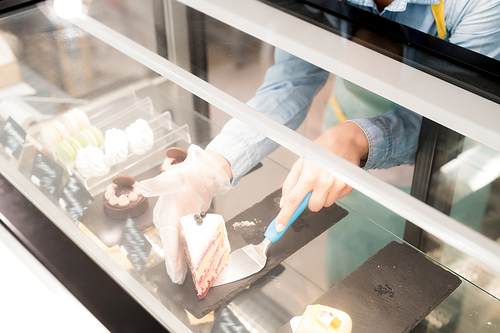 High angle close up of unrecognizable young woman taking piece of strawberry cake from window display and offering in to customers in small cafe, copy space