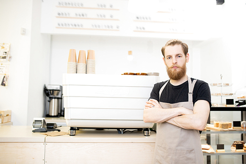 Serious handsome young entrepreneur in apron standing at espresso machine and  while crossing arms on chest in coffee shop