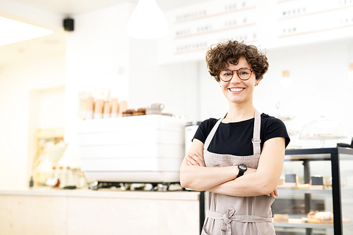 Smiling attractive hipster female barista standing in coffee shop and crossing arms on chest while 