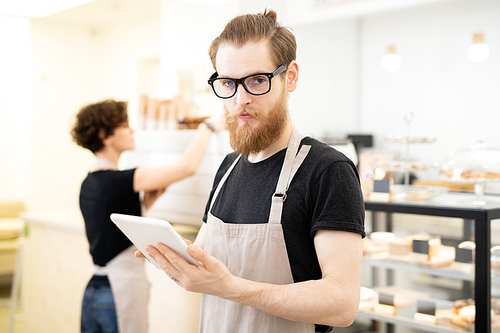 Serious handsome hipster young bearded waiter in glasses standing in bakery store and  while using digital tablet
