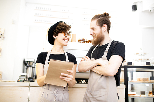 Positive excited coffee shop colleagues in aprons standing at counter with espresso machine and discussing delivery of goods while viewing notes in sketchpad