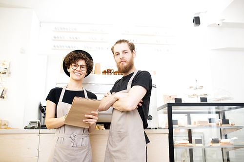 Content confident team of coffee shop: smiling young man and woman in aprons standing at counter and  while analyzing notes in sketchpad