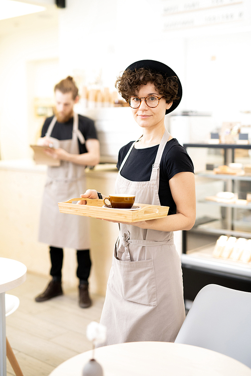 Content attractive hipster lady waitress in apron carrying wooden tray with coffee cup and  in coffee shop