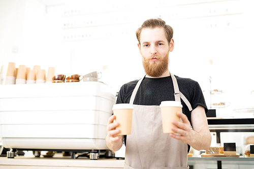 Content confident barista man in apron standing in coffee shop and  while giving takeout coffee cups to customer