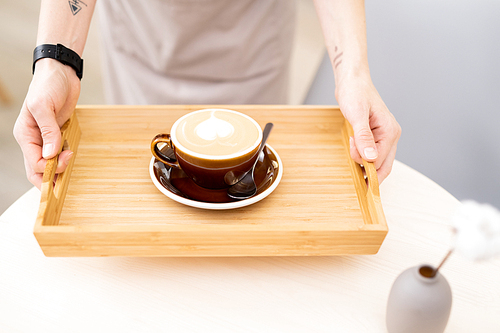 Close-up of morning cappuccino with heart drawn on steam placed on wooden tray, waitress bringing coffee