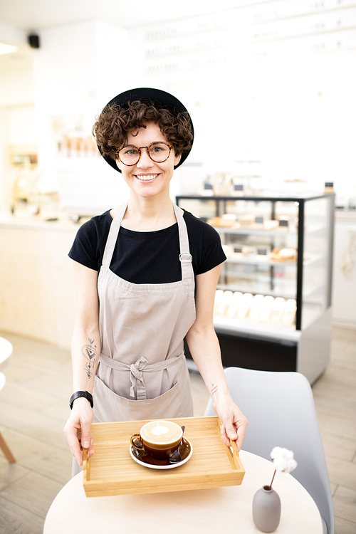 Happy attractive hipster woman in apron and black hat holding wooden tray with cappuccino and  cheerfully, she working in coffee shop
