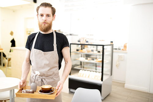 Serious handsome young barista man carrying tray with cappuccino and chocolate doughnut in modern bakery shop and