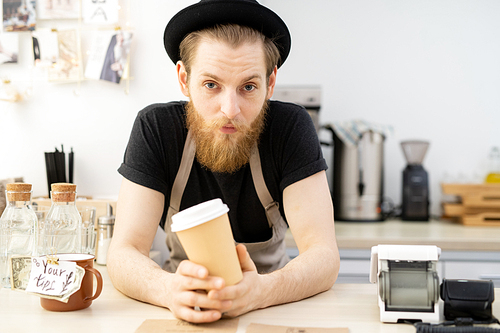 Content frowning handsome barista man in black hat leaning on counter and holding takeout coffee cup while  in coffee house
