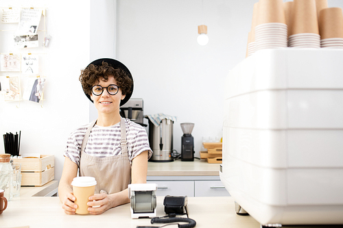 Smiling positive curly-haired hipster barista woman in hat standing at counter with espresso machine and  while holding disposable coffee cup