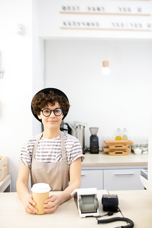 Content beautiful lady in hat and apron standing at counter with payment terminal and coffee cup in coffee house and 