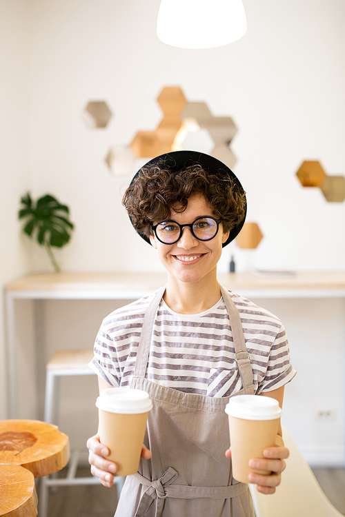 Cheerful attractive hipster barista in hat and glasses standing in modern coffee shop and  while offering to drink coffee
