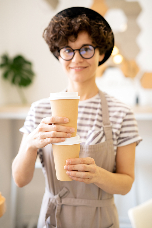 Positive beautiful curly-haired young lady in apron and hat holding two disposable coffee cups and , barista brewing tasty coffee in cafe