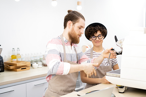 Content experienced handsome bearded barista in apron teaching new employee to use espresso machine, he explaining how to steam milk