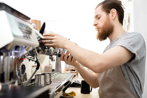 Serious busy handsome barista with beard wearing apron preparing espresso machine for work and attaching portafilter