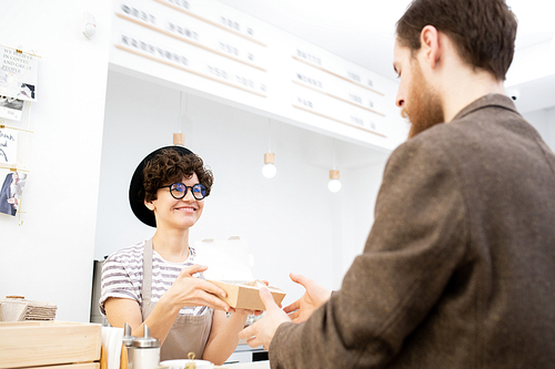 Smiling attractive young female barista in black hat and eyeglasses standing behind counter and giving paper box with sweets to customer in cafe