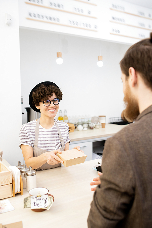 Hipster man with beard buying snack in coffee shop, cheerful positive young woman in black hat giving paper box to customer