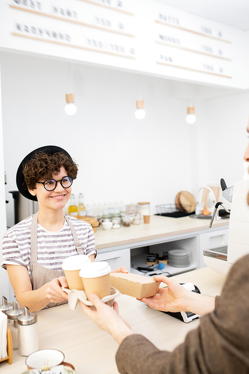 Coffee to go: positive attractive young hipster lady in black hat giving takeout coffee cups and small cardboard box to customer in coffee shop