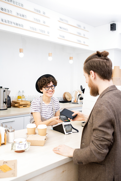 Hipster bearded guy in casual jacket putting smartphone to terminal while paying for coffee to go in cafe, positive barista smiling at customer