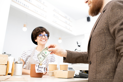 Close-up of bearded man in jacket leaving tip for friendly barista while buying coffee and snacks in coffee shop
