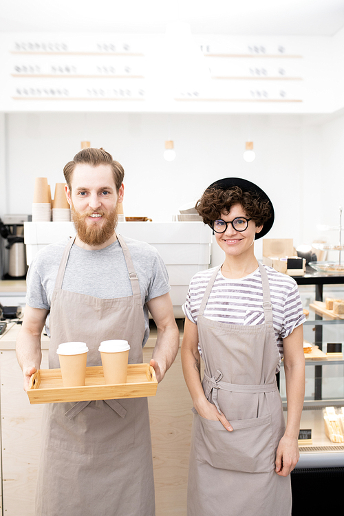 Cheerful positive young baristas in aprons working in modern coffee shop: they , smiling man holding wooden tray with coffee cups