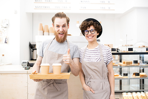 Barista brewing excellent coffee: cheerful excited bearded barista man holding tray with takeout coffee cups and showing thumb-up, pretty lady in apron 