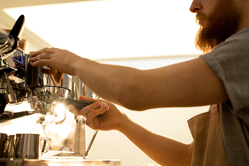 Close-up of serious busy bearded barista man in apron attaching portafilter to espresso machine while brewing coffee in cafe