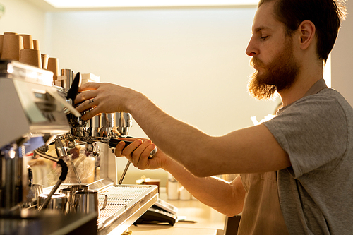 Serious handsome bearded barista in apron standing at counter and using espresso machine while preparing coffee in restaurant