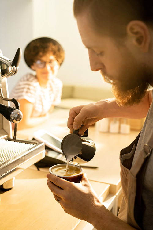 Serious concentrated hipster bearded barista pouring milk into coffee cup while making cappuccino for customer in coffee shop