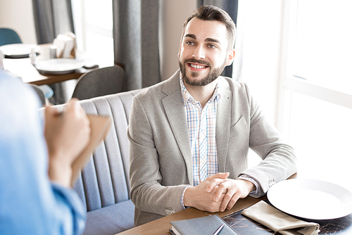 Happy positive handsome young businessman sitting at table with plate and smiling to waitress while making order in restaurant