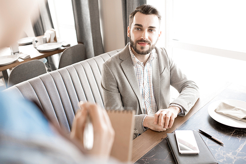 Content confident bearded young businessman in casual jacket sitting at restaurant table and talking to waitress while knowing menu of business lunch