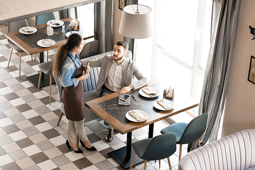 Directly above view of qualified waitress in apron talking to handsome customer in casual jacket while answering his question about food in restaurant