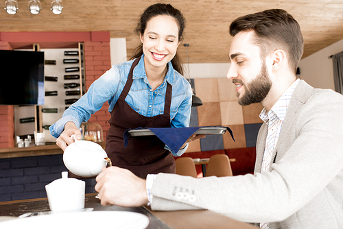 Positive careful beautiful waitress in apron holding tray with napkin and pouring hot tea into cup to smiling customer while serving him in restaurant