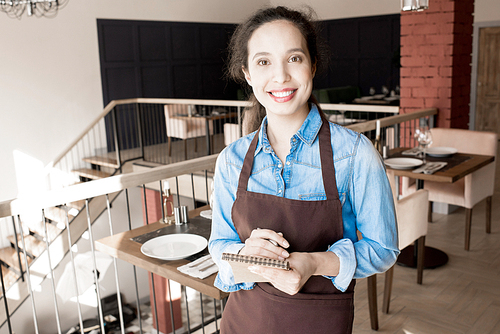 Portrait of cheerful excited beautiful young mixed race waitress in apron holding pen and notepad for writing orders and standing in modern restaurant room