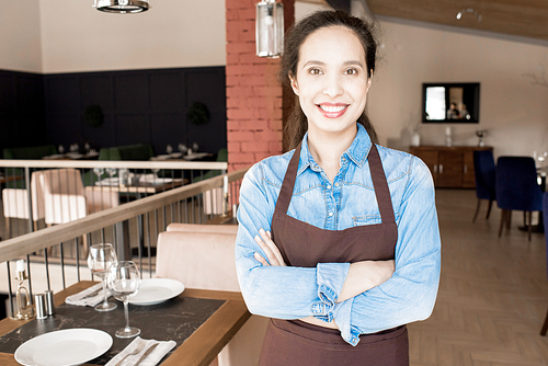 Positive optimistic attractive young mixed race waitress in apron crossing arms on chest and smiling at camera while standing in spacious restaurant room